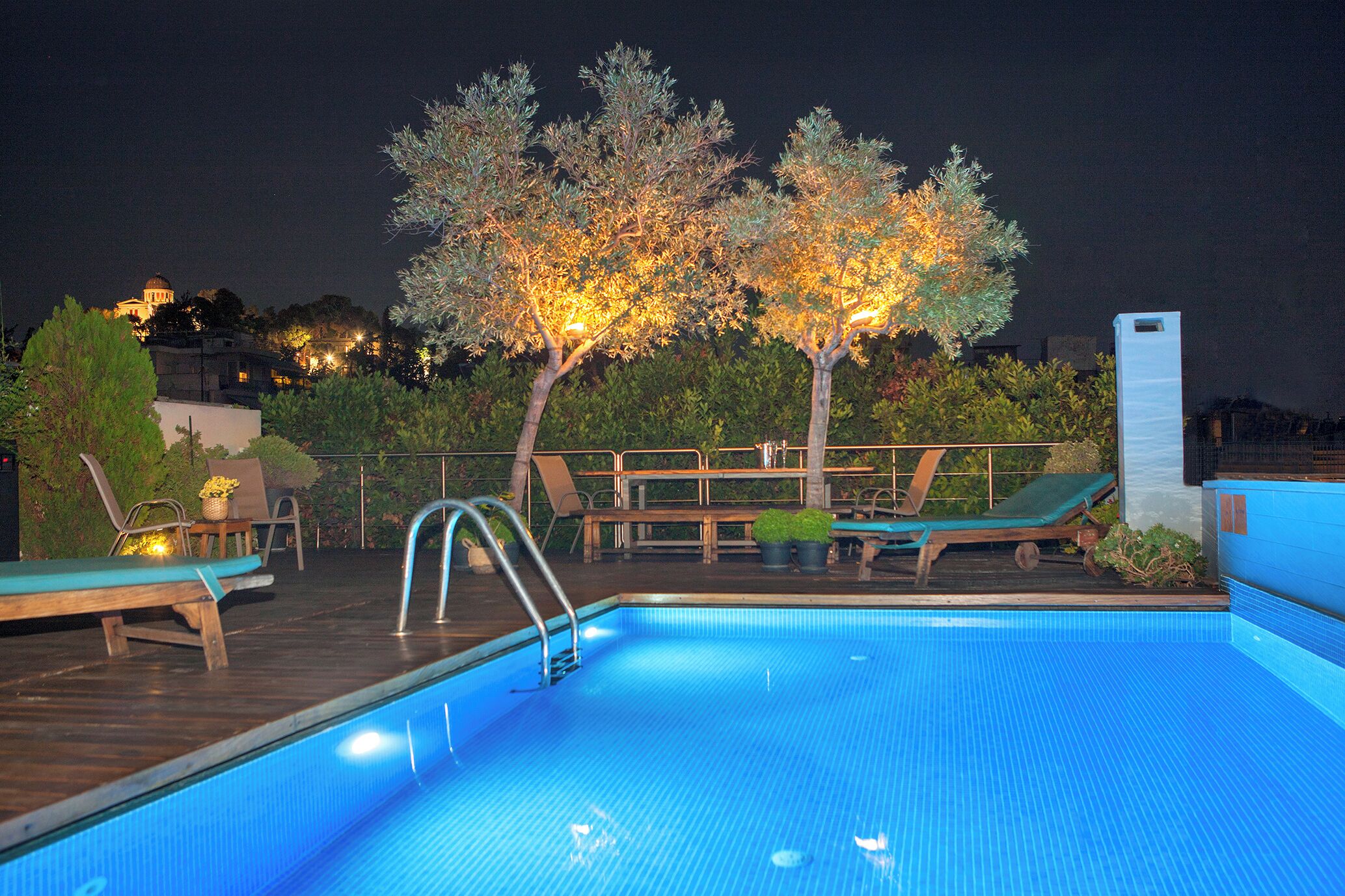 A swimming pool with lounge chairs and a wooden deck, surrounded by trees and a view of a distant building.