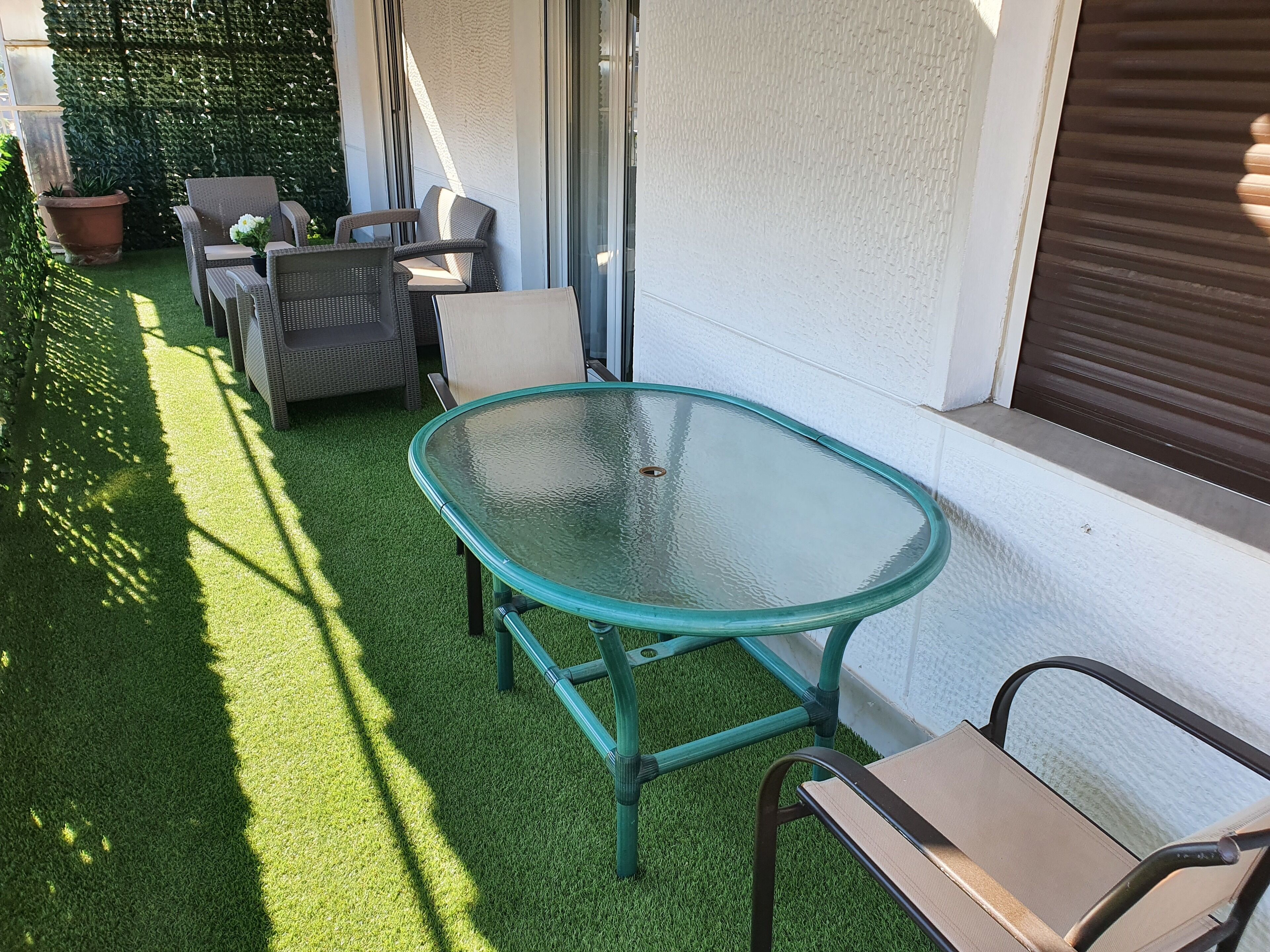 Uma área externa com espaço para sentar, incluindo uma mesa e cadeiras de vidro, cercada por grama sintética verde e uma parede branca.