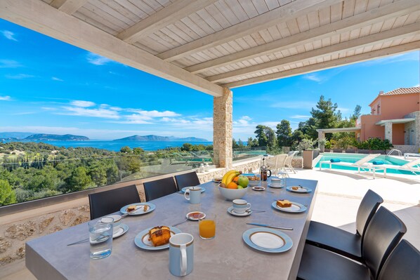 Una mesa de comedor con platos, cubiertos y una cesta de frutas, con vista a una piscina y un paisaje pintoresco.