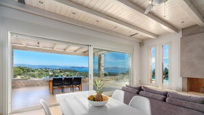A modern dining area with a table, chairs, and a bowl of fruit. There is a view of the outdoors through large glass doors. - Villa Emmelia & Guesthouse Amazing View With Pool (Πόρτο Χέλι)
