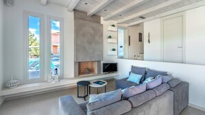 A modern living room with a grey sofa, a fireplace, a TV, and a view of a pool. - Villa Emmelia & Guesthouse Amazing View With Pool (Πόρτο Χέλι)