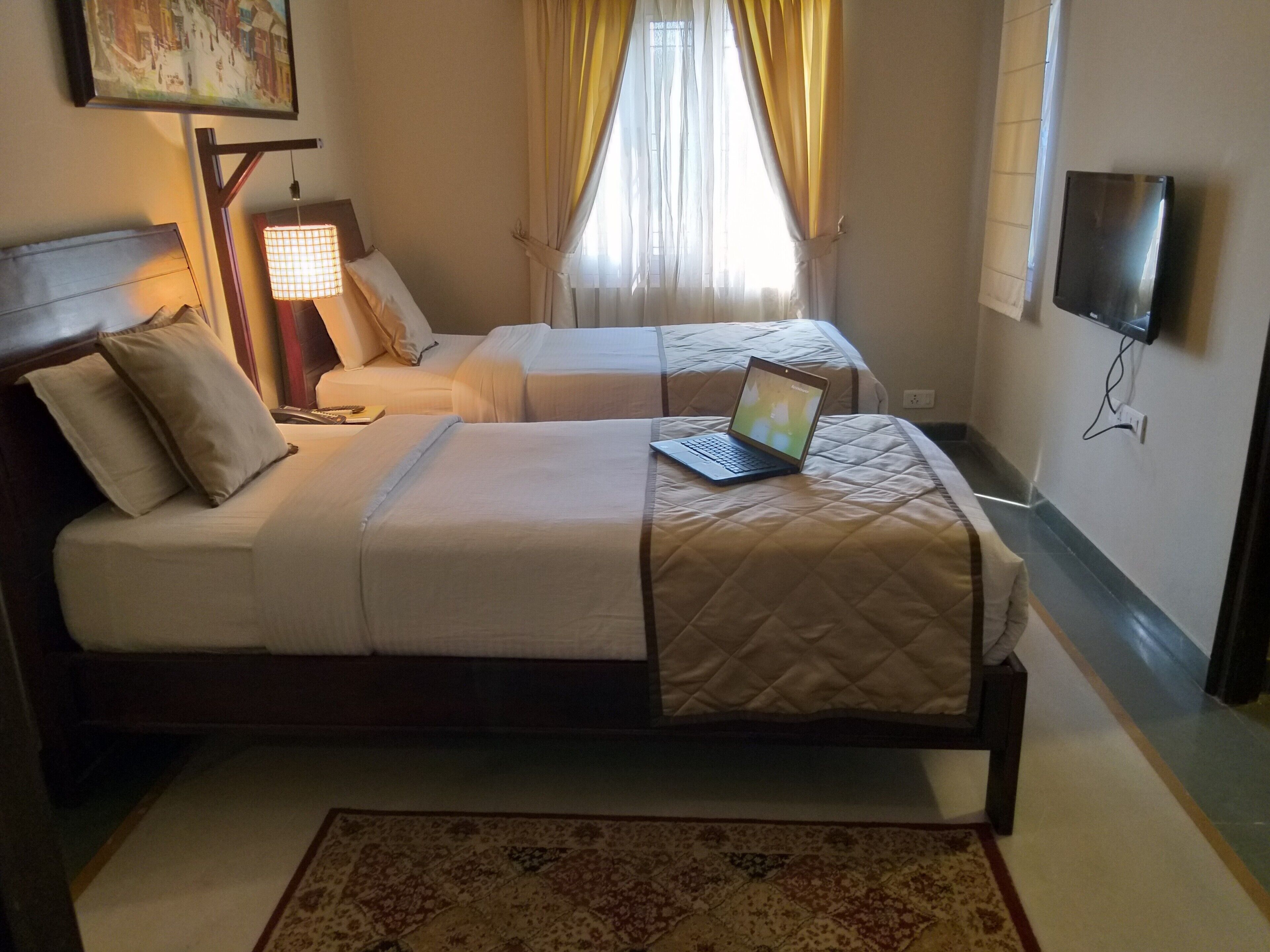 A hotel room with two beds, a laptop on the right bed, a TV on the wall, and a painting above the left bed.
