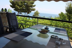 Een glazen tafel met een schaal lekkernijen, een paar brillen en een kopje op een balkon met uitzicht op zee en groen.