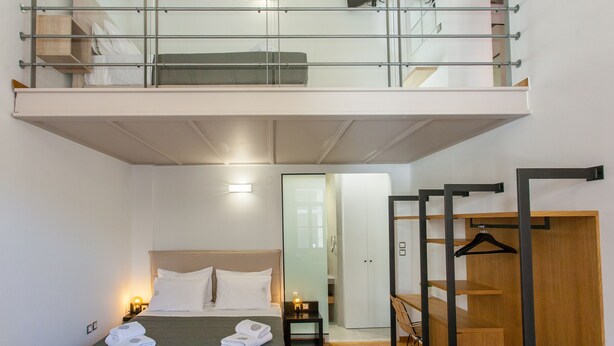 A compact hotel room with a lofted sleeping area, a bed with white linens, a small bedside table with a lamp, and a wooden wardrobe. - Agora Suites  - Stay in the HEART of CHANIA (Χανιά)