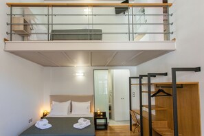 A compact hotel room with a lofted sleeping area, a bed with white linens, a small bedside table with a lamp, and a wooden wardrobe. - Agora Suites - Stay in the HEART of CHANIA (Χανιά)