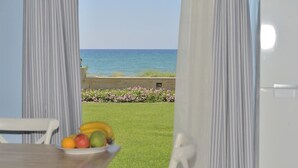 Vue depuis une pièce où l’on aperçoit une table et un saladier rempli de fruits, donnant sur un espace verdoyant parsemé de fleurs et bordé d’une étendue d’eau.