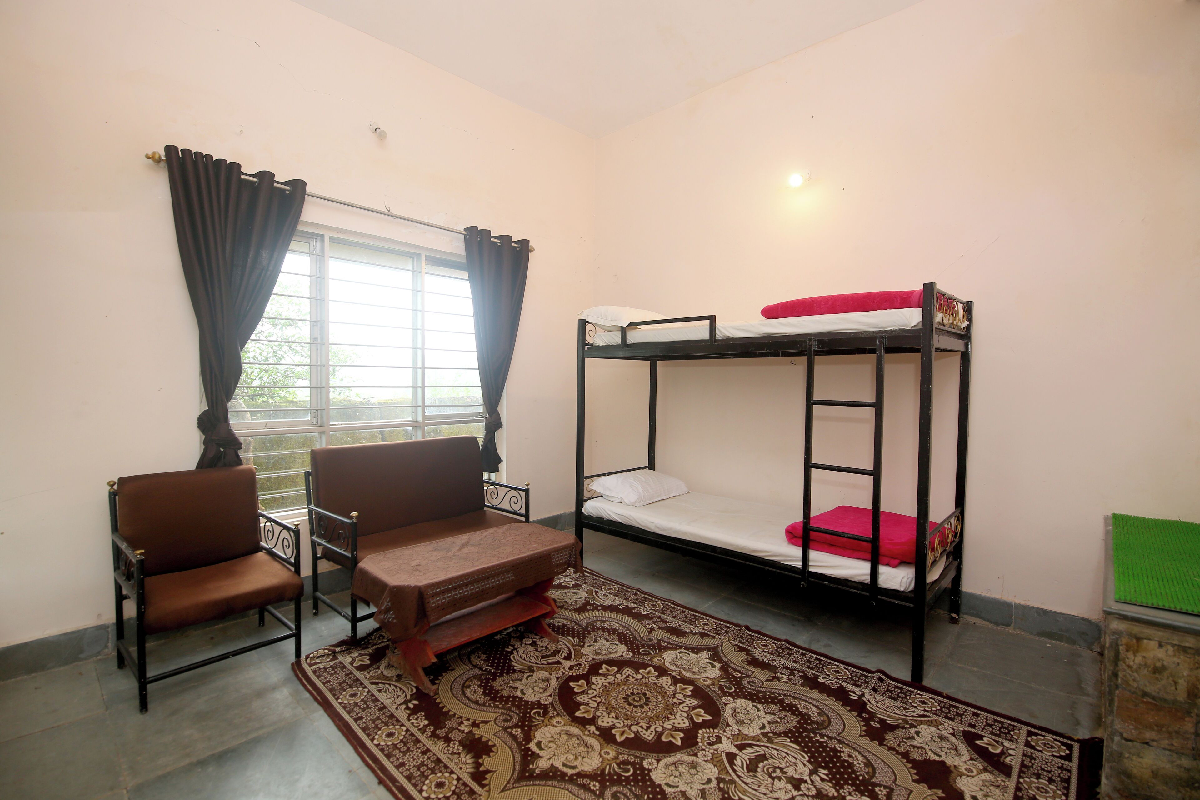 A dormitory room with a bunk bed, two chairs, a patterned rug, and a window with curtains.