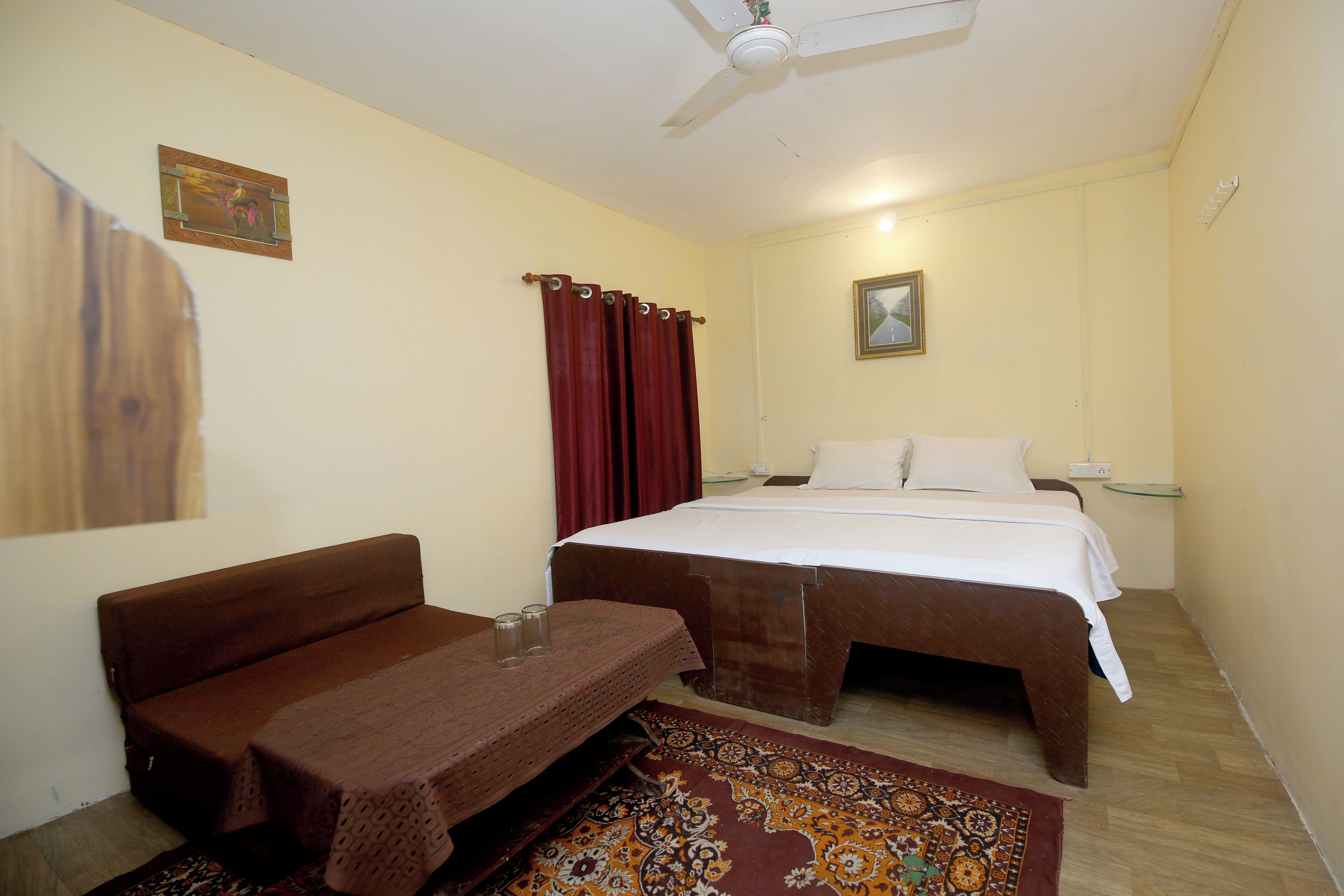 A bedroom with a bed, a sofa, a small table, a ceiling fan, and a patterned rug.