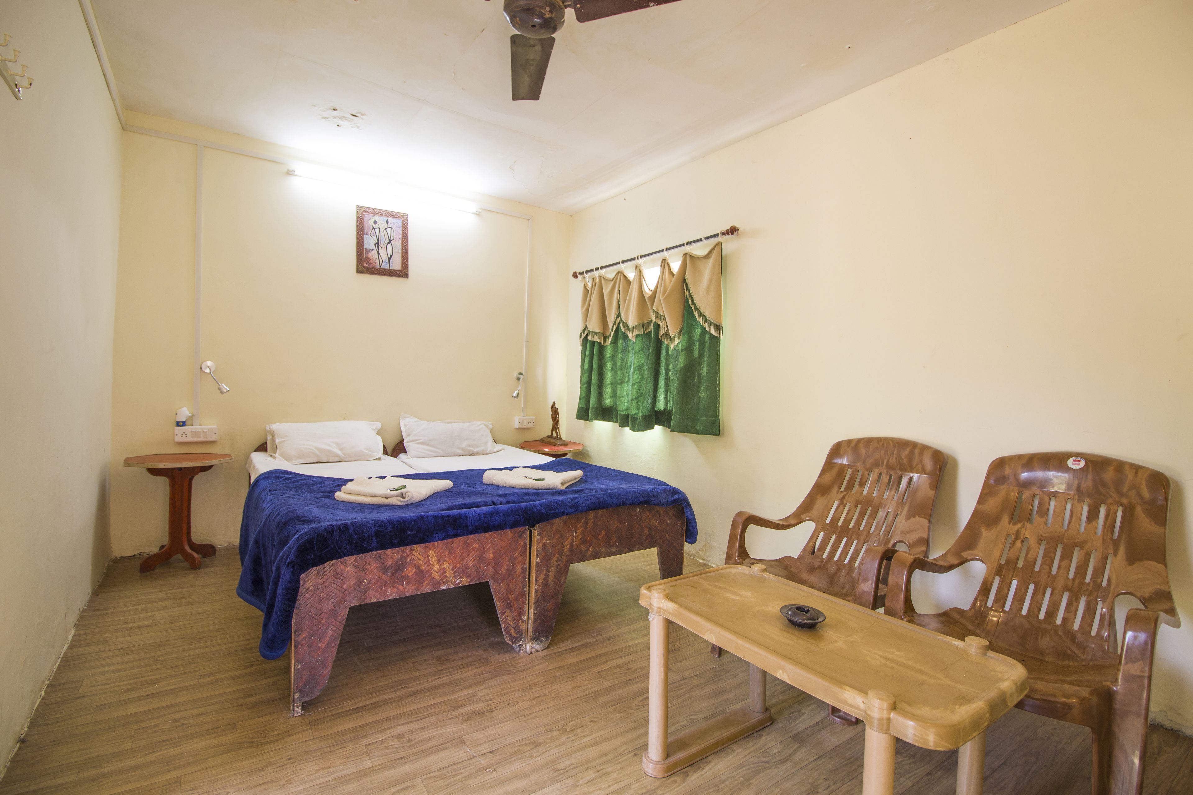 Une pièce comprenant un lit, deux chaises, une petite table, une table de chevet, un ventilateur de plafond et un rideau vert.