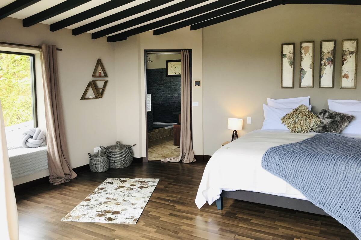Une chambre avec un plancher en bois, un lit avec une literie blanche, une couverture bleue et une table de chevet avec une lampe. Il y a une fenêtre avec des rideaux, une étagère murale avec des objets décoratifs et une porte qui mène à une autre pièce.