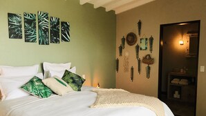 Un lit bien fait, avec du linge de lit blanc et des coussins à motifs verts, un mur orné d’œuvres d’art représentant des feuilles tropicales, et une porte donnant sur une salle de bain où se trouvent des serviettes.