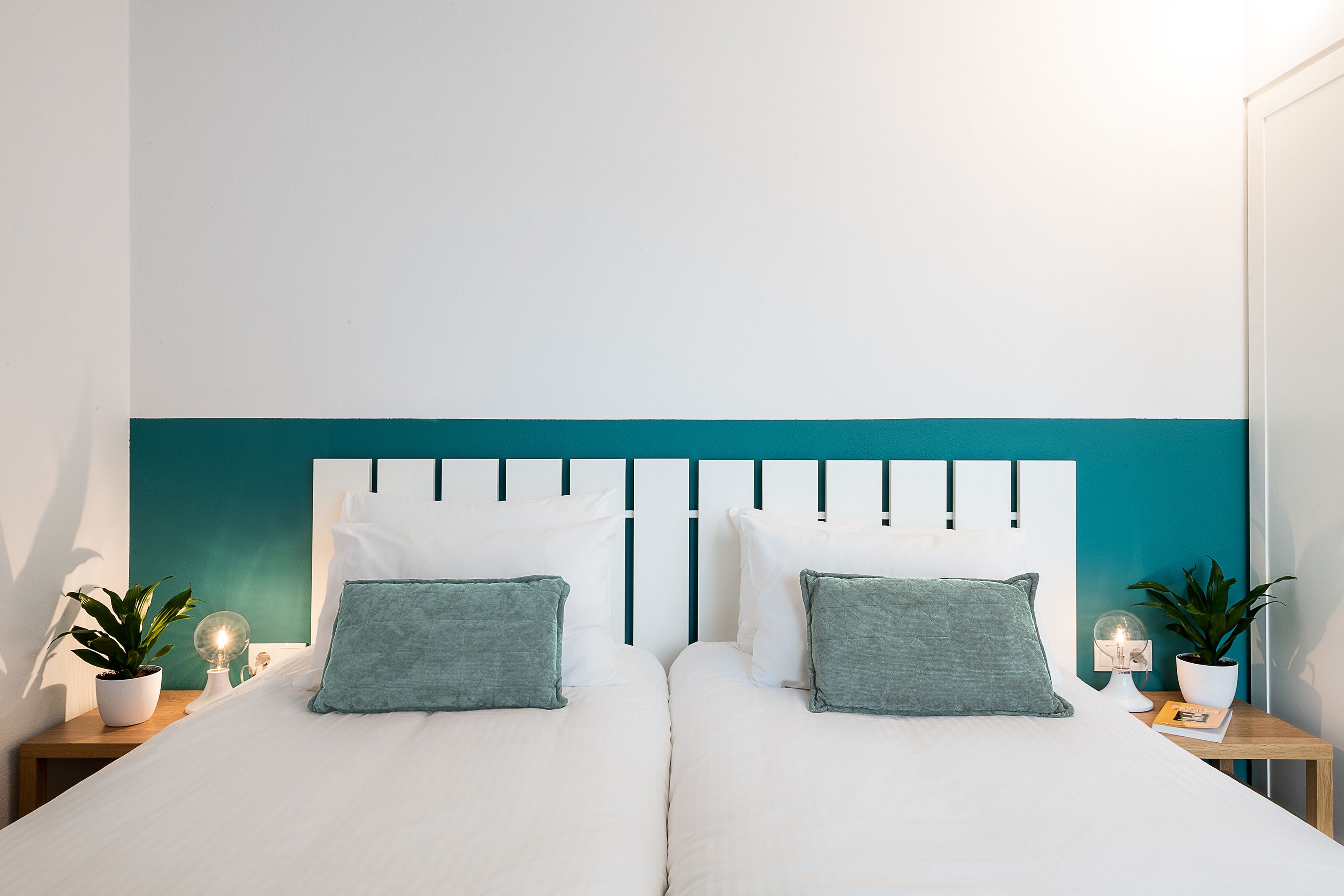 A double bed with white bedding and teal headboard, flanked by bedside tables with potted plants and a globe lamp.