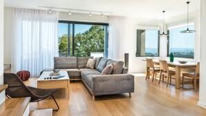 Un salon moderne avec un canapé d’angle, une table à manger avec des chaises et de grandes fenêtres donnant sur l’extérieur.