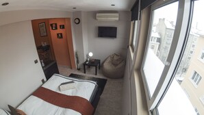 A hotel room with a bed, a bedside table with a lamp, a bean bag chair, a wall-mounted TV, and a window with a view of buildings.