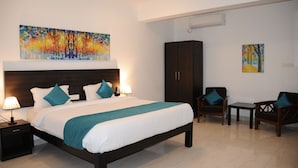 A hotel room with a large bed, two bedside tables with lamps, a dark wooden wardrobe, a small table with chairs, and two paintings on the walls. - JW Heritage (Anjuna)