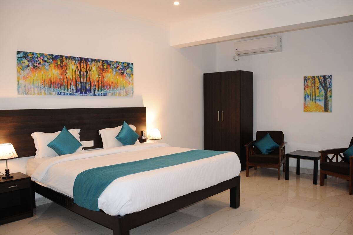 Habitación de hotel con una cama grande, dos mesitas de noche con lámparas, un armario de madera oscura, una pequeña mesa con sillas y dos cuadros en las paredes.
