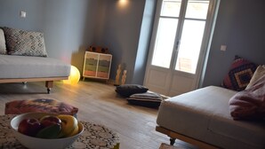 Een knus ingerichte kamer met een bed, een bank, een klein tafeltje met een fruitschaal en een raam met natuurlijk licht.