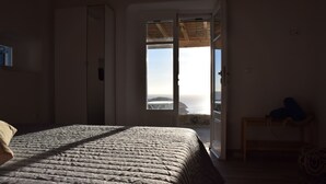 Een slaapkamer met een bed, een kledingkast, een bankje en een deur die uitkomt op een balkon met uitzicht op zee.