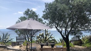 A wooden bench and table under a large umbrella, surrounded by plants and a fence, with a view of the sea and hills in the background. - Beachfront family house (Ουρανούπολη)