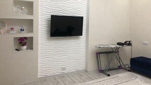 Un soggiorno con una TV a schermo piatto montata su una parete strutturata, un piccolo asse da stiro con ferro da stiro e un divano blu.