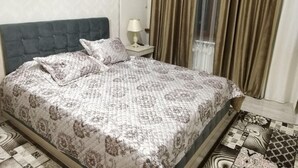Un letto rifatto con cura, con copripiumino e cuscini decorati, un comodino con una lampada e una finestra con tende.