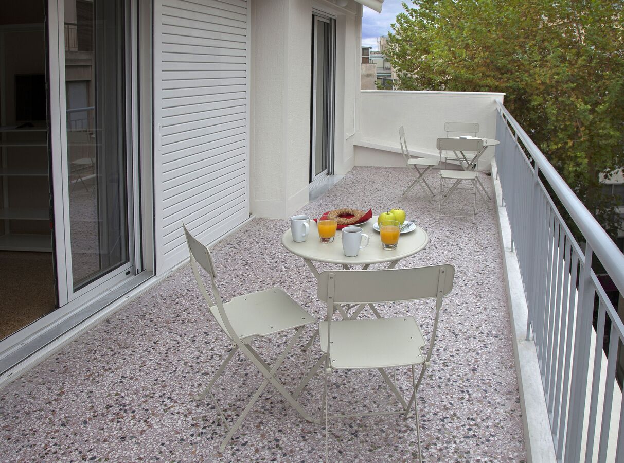 A balcony with a table set for two, chairs, and a small round table with a fruit bowl and drinks.