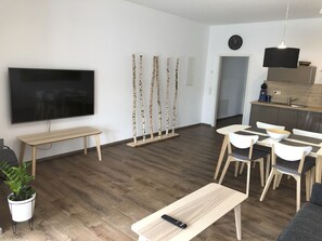 A modern living room with a flat-screen TV, a wooden bench, birch tree sculptures, a dining area with a white table and chairs, and a kitchen area with a sink and countertop.