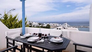 Une table dressée pour deux personnes, avec des assiettes, des verres et une bouteille, donnant sur une ville côtière aux bâtiments blancs et sur la mer.