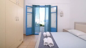Une chambre à coucher avec un lit, deux serviettes, une table de chevet, une lampe fixée au mur et une vue sur l’extérieur, visible à travers une porte ornée de rideaux bleus.