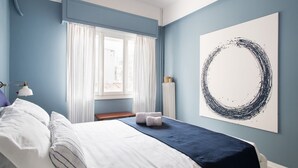Une chambre à coucher avec un lit, une fenêtre avec des rideaux, une œuvre d’art accrochée au mur et une lampe de chevet.
