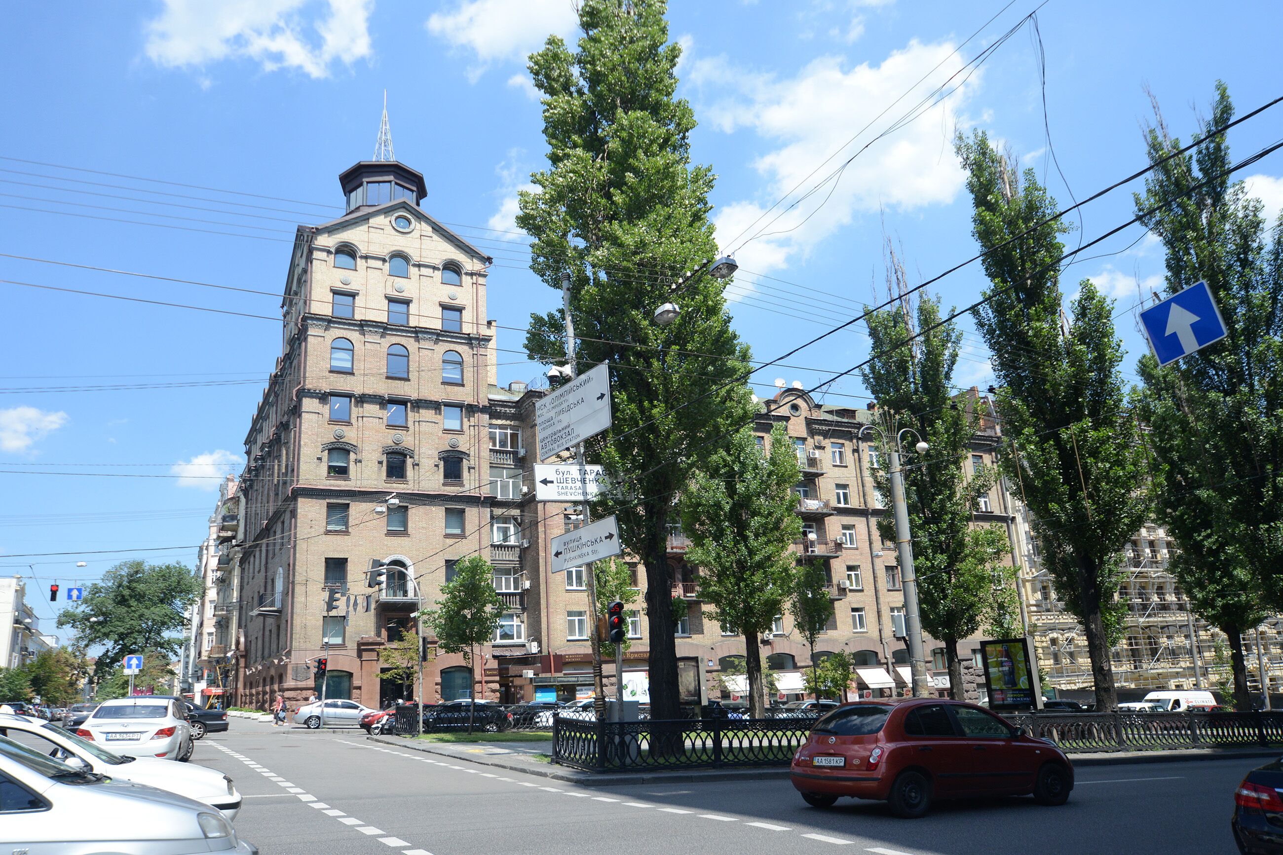 A multi-story building with a prominent corner tower, surrounded by trees and parked cars.