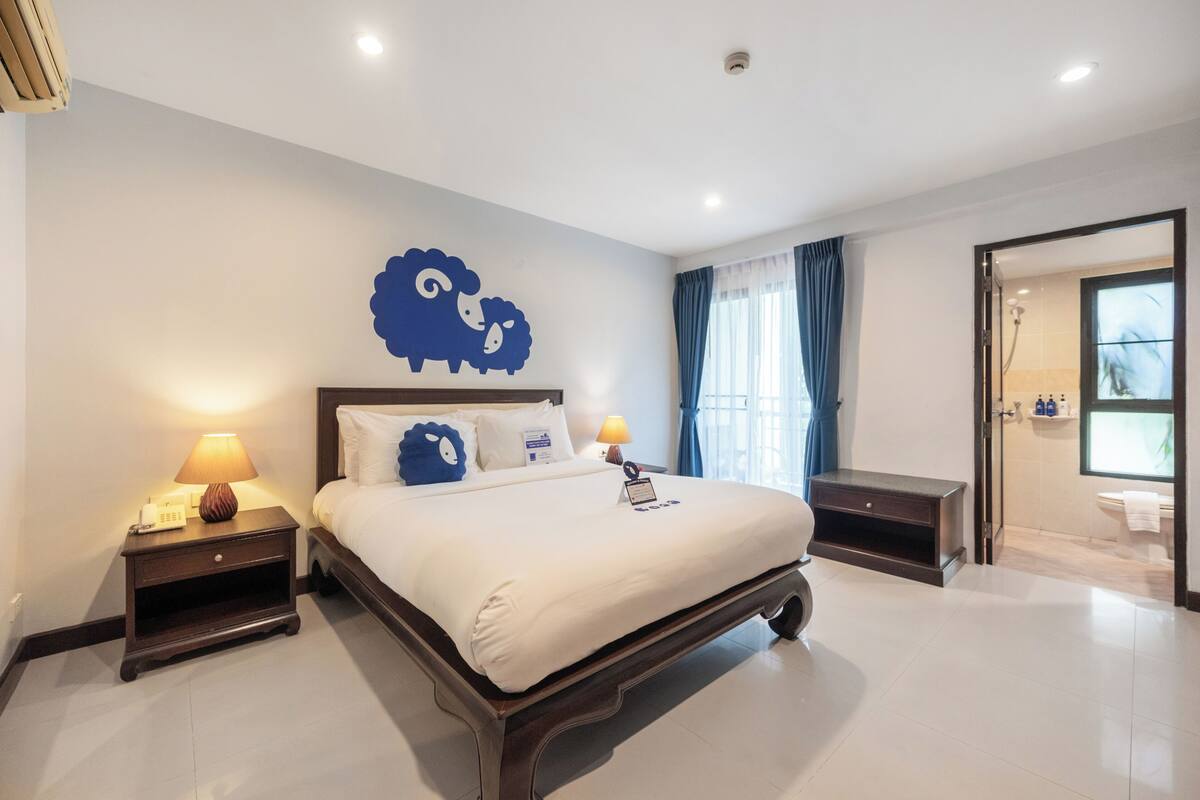 A hotel room with a bed, bedside lamps, a wall decoration, a bathroom visible through an open door, and a window with curtains.