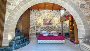 Un dormitorio con pared de piedra, una cama con manta roja, una escalera de madera y un vidrio que da a un armario.