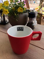 Una taza roja con una etiqueta que dice "NAP HOTEL - CAFÉ RESTAURANT" sobre una mesa de madera.