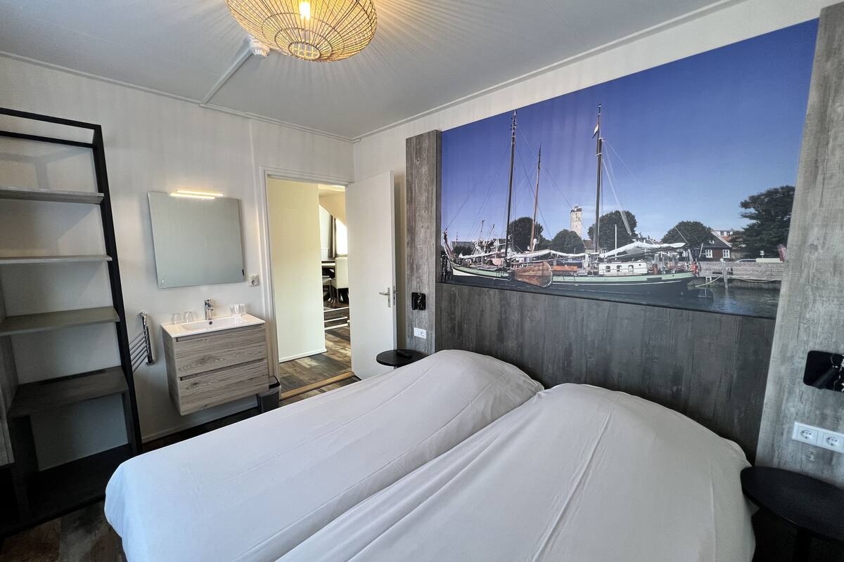 A bedroom with a large bed, a bathroom with a mirror and sink, and a view of boats through a window.