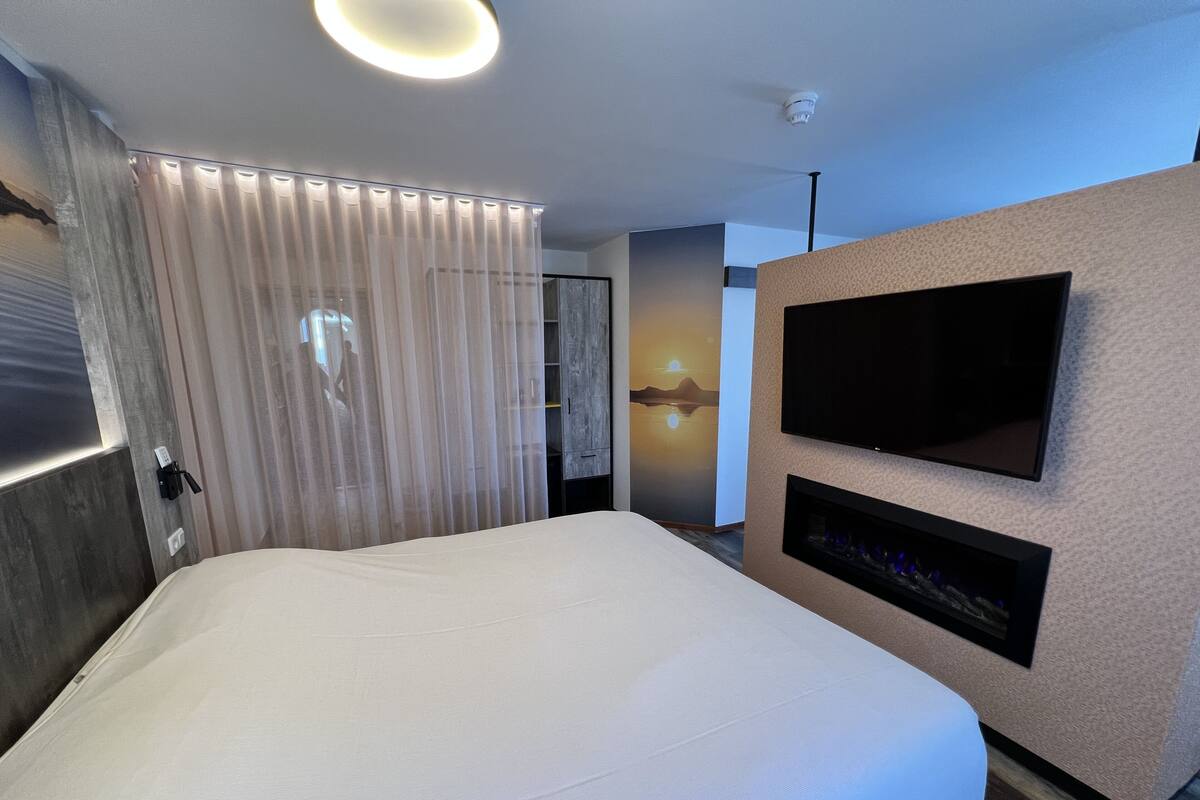 A modern hotel room with a large bed, a flat-screen TV mounted on the wall, and a built-in fireplace.