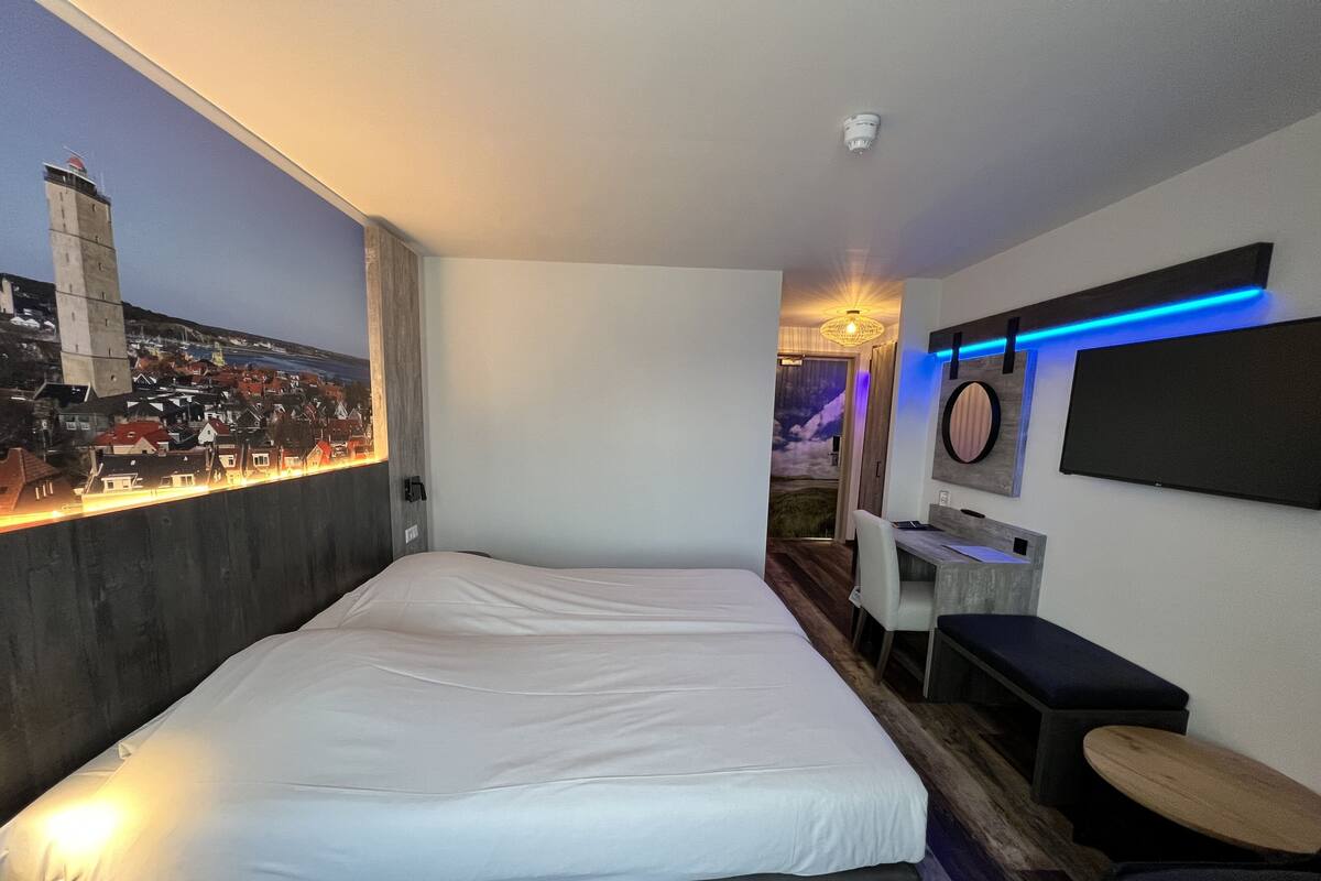 A modern hotel room with a large bed, a desk, and a cityscape mural on the wall.