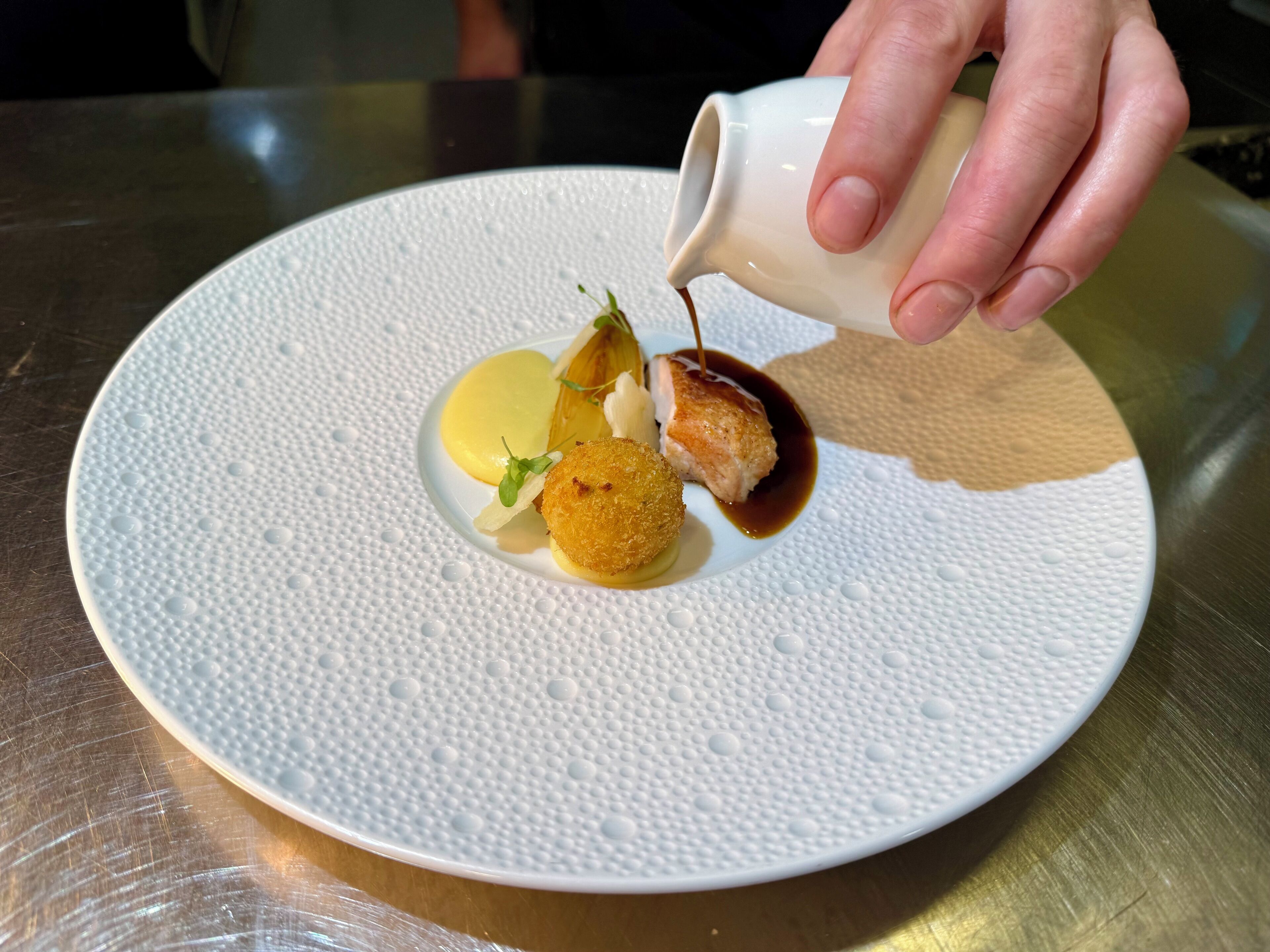 A white plate with a textured surface, a piece of food, and a hand pouring a dark liquid onto it.
