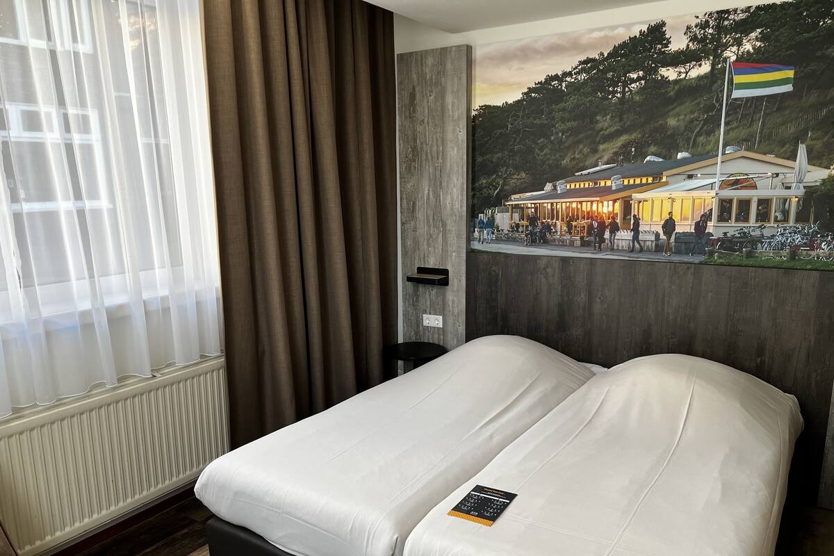 A hotel room with a bed, a window with curtains, a radiator, and a wall-mounted picture.