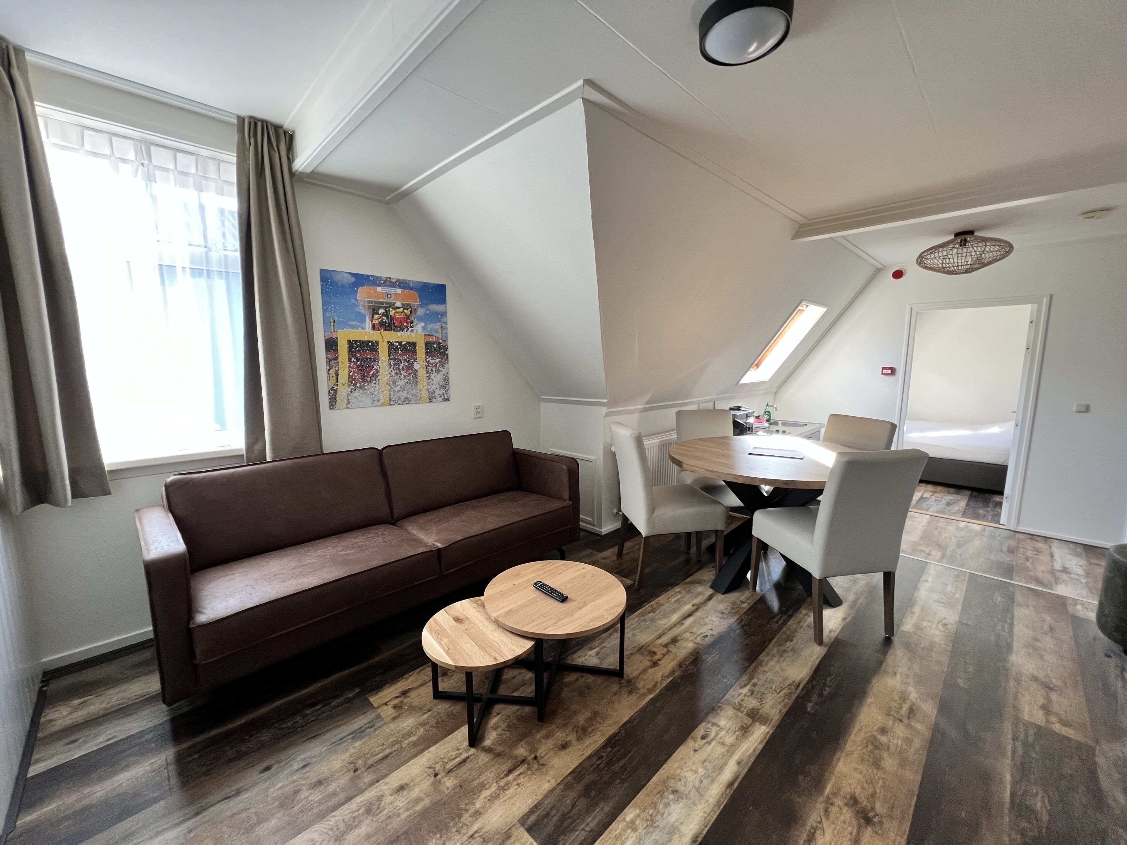 Un salon moderne avec un canapé marron, deux petites tables rondes en bois, une table à manger avec des chaises, un coin cuisine et une chambre avec un lit et une fenêtre de toit.