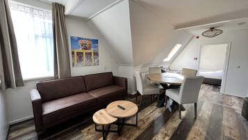 Una sala moderna con un sofá marrón, dos mesas redondas de madera, una mesa de comedor con sillas, una zona de cocina y un dormitorio con cama y tragaluz.