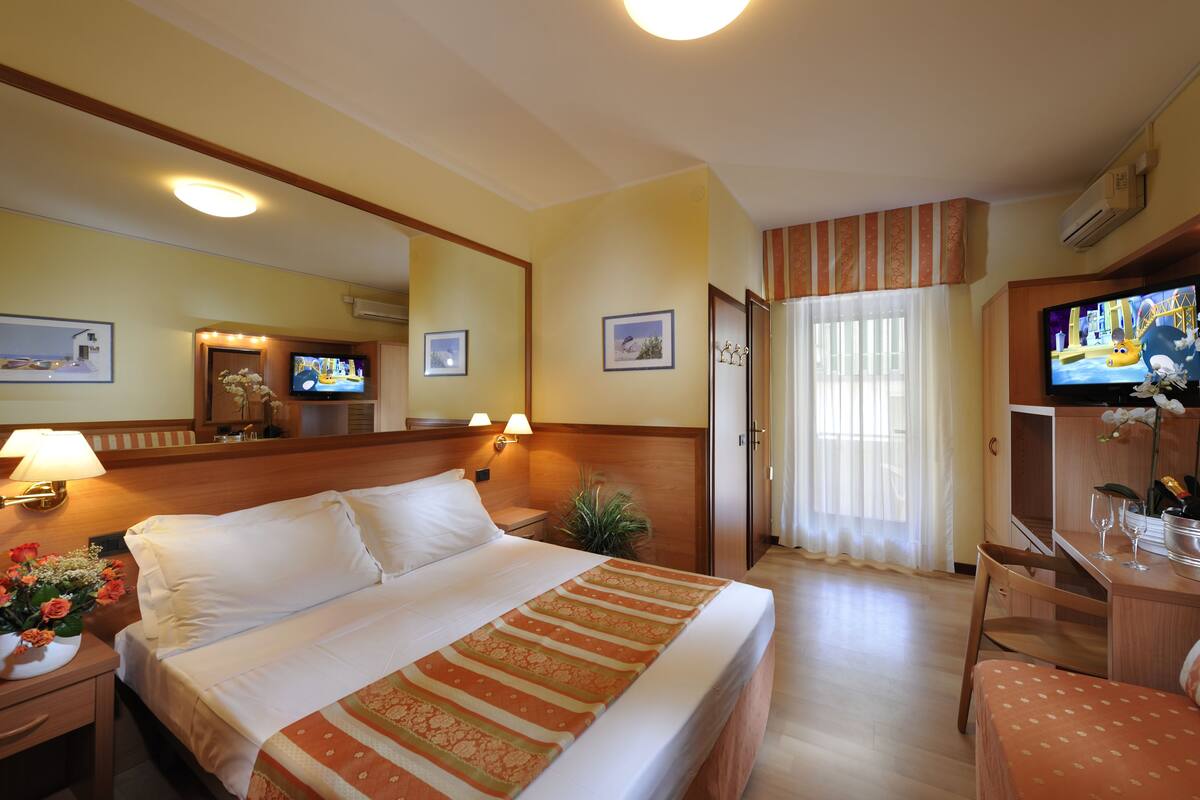 Une chambre d’hôtel comprenant un lit, une table de chevet avec une lampe, un téléviseur, un bureau avec une chaise et une fenêtre avec des rideaux.