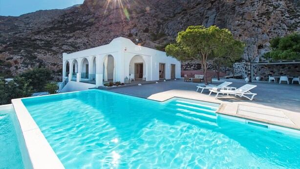 A swimming pool with a white building in the background, surrounded by mountains. - Villa Adrali, 4 bedrooms, private pool (ΘΗΡΑ)