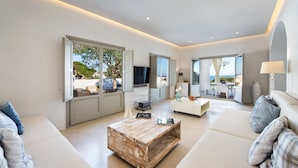 A modern living room with a large window, a wooden coffee table, and a white sofa. - Villa Adrali, 4 bedrooms, private pool (ΘΗΡΑ)