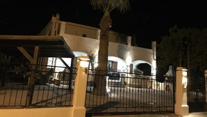 A gated entrance with a palm tree, a white building with arches, and a well-lit pathway. - Welcome To Our Traditional Experience House (Ρέθυμνο)