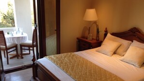 A neatly made bed with a wooden headboard, a bedside table with a lamp, and a small table with chairs outside the room. - Welcome To Our Traditional Experience House (Ρέθυμνο)