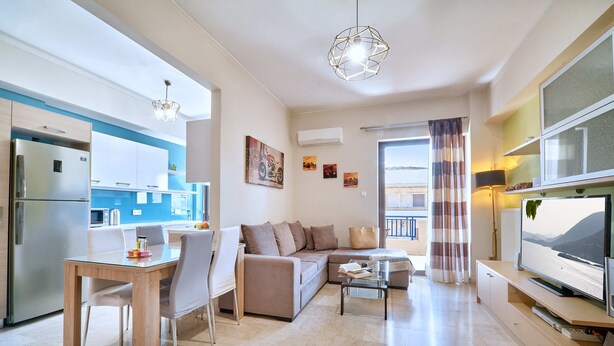 A modern living room with a sofa, coffee table, and TV. There is a kitchen area with a refrigerator and a dining table with chairs. - Modern Apartment, just 70 meters from the sea! (Ρέθυμνο)