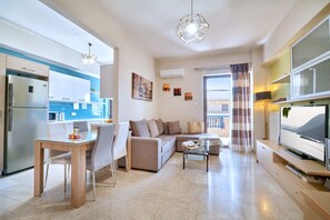 A modern living room with a sofa, coffee table, and TV. There is a kitchen area with a refrigerator and a dining table with chairs. - Modern Apartment, just 70 meters from the sea! (Ρέθυμνο)