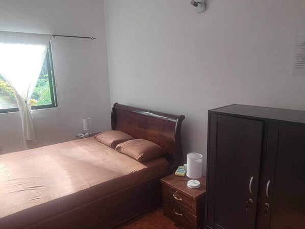 A bedroom with a bed, a nightstand, a wardrobe, a window with curtains, and a wall-mounted light fixture. - Papaya Guesthouse (Mahé Island)