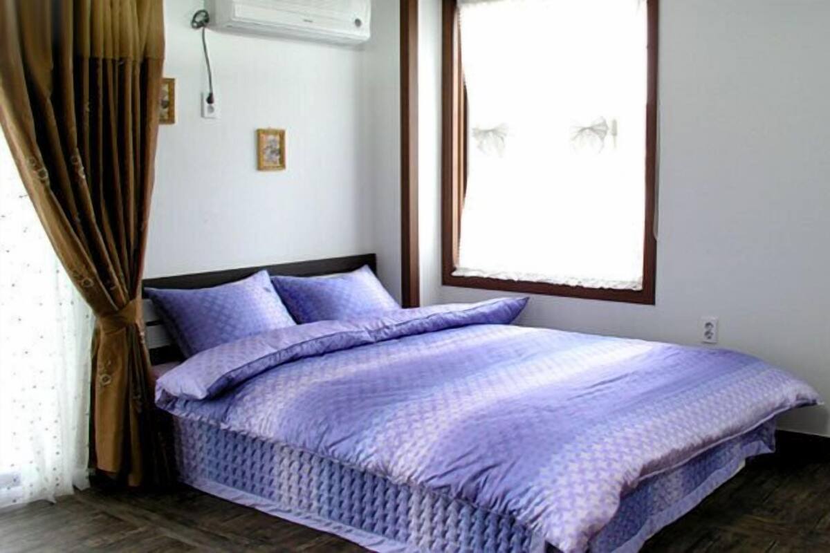 Een slaapkamer met een bed, gordijnen, een raam en een luchtverkoeler aan de muur.